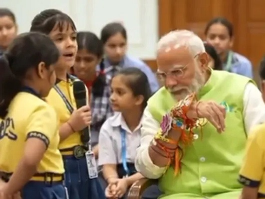 "प्रधानमंत्री नरेंद्र मोदी के हाथ पर रंग-बिरंगी राखियां सजी हुई हैं, जबकि वे स्कूल के बच्चों के साथ रक्षाबंधन का त्योहार मना रहे हैं।"